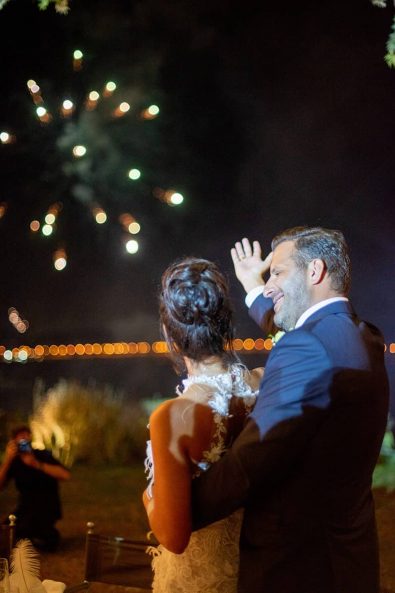 Bride in groom's arms watching the firework show at their wedding party reception.