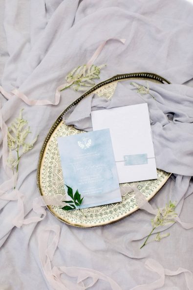 Fine art photography of wedding invitation in light blue and white shades and silk ribbons on a gold serving tray.