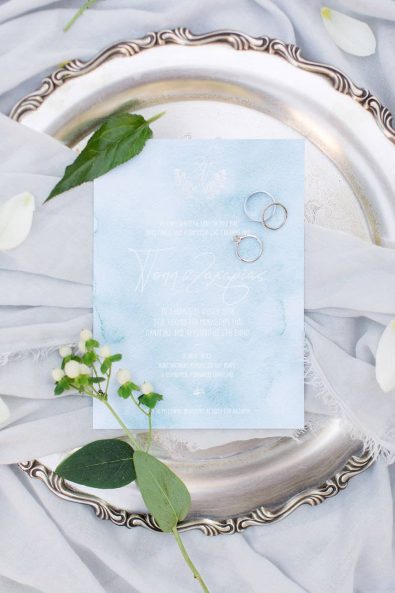 Detail shot of watercolor light blue wedding invitation and white gold wedding rings on an aged silver serving tray for a destination wedding in Greece.