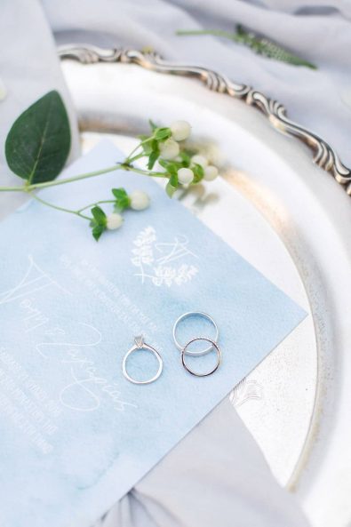 Close up shot of the white gold and diamond engagement ring and wedding bands on a light blue watercolor paper wedding invitation.