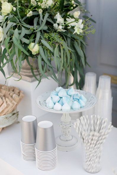 Refreshments stand with white and green flower arrangement, glass serving tray with light blue meringues and silver paper cups for flavored water.