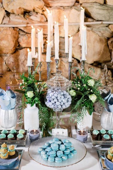 Close up shot of summer wedding candy bar in blue shades, with marbled macarons, cupcakes with light blue buttercream frosting, religieuse french desserts, and sugar-coated almond koufeta, decorated with white candles and flower arrangement with greenery and roses.