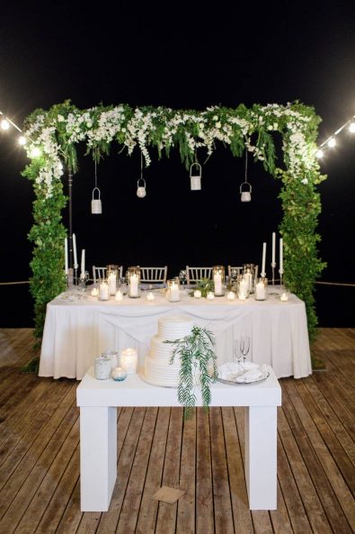 Shot of table with the elegant and simple wedding cake in front of the bridal table with the white flower and greenery decorated canopy.