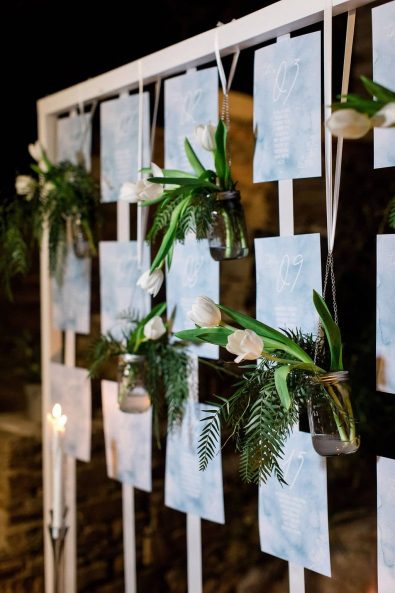 Creative backdrop inspiration with wedding stationery glued in satin ribbons and hanging glass jars with white tulips and greenery.